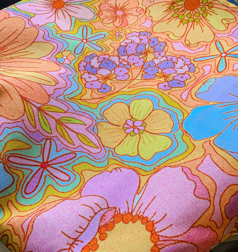 May include: A close-up of a fabric with a vibrant floral pattern. The design features large, stylized flowers in shades of orange, pink, yellow, and blue. The background has a wavy, abstract design in complementary colors, creating a retro, psychedelic aesthetic.
