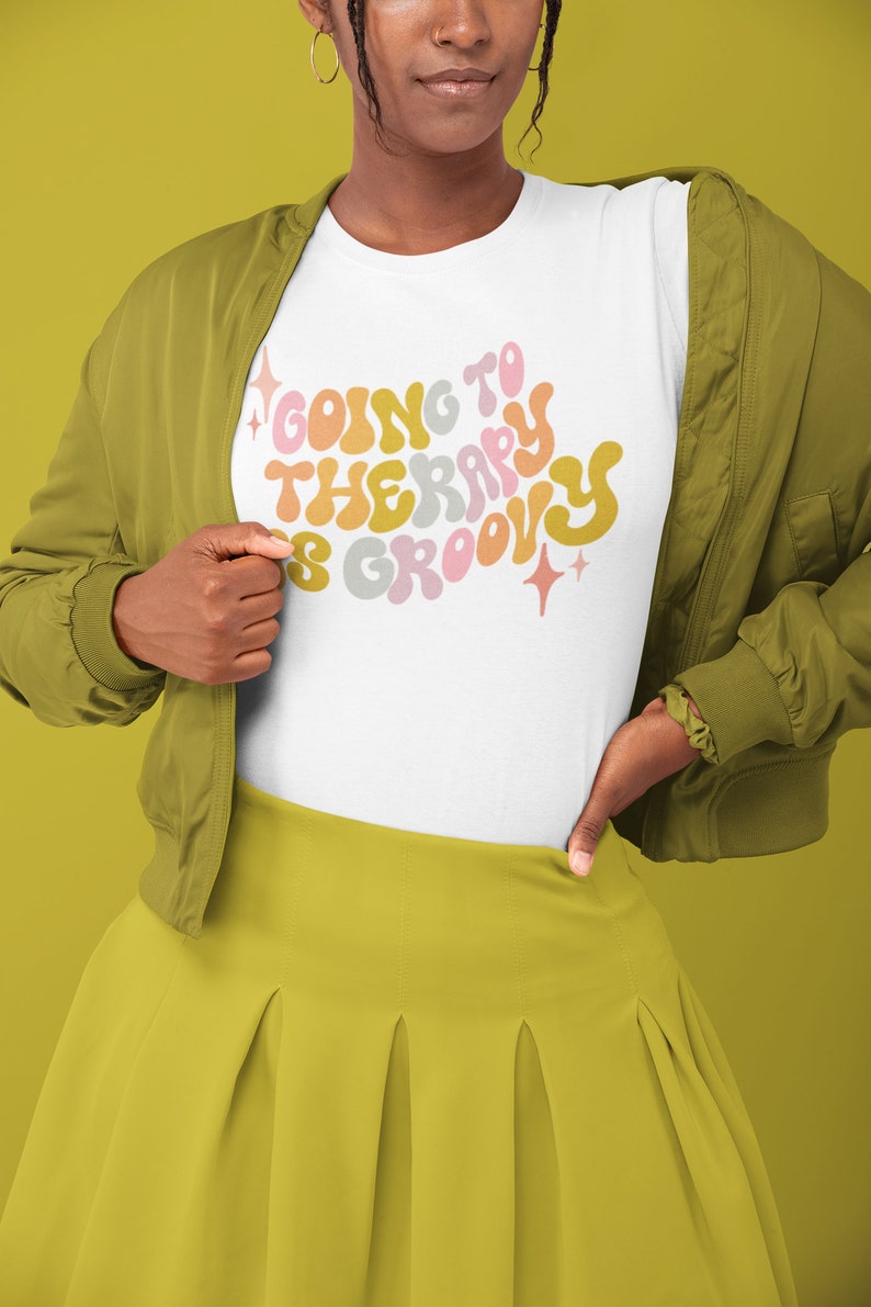 Puede incluir: Una camiseta blanca con un gr&aacute;fico de estilo retro que dice "Ir a terapia es genial" en letras coloridas y onduladas. La camiseta la lleva puesta una persona con una chaqueta verde y una falda amarilla.