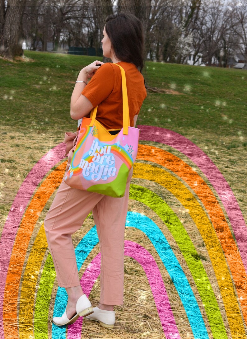 May include: A colorful tote bag with yellow straps and the words "You are magic" printed on the front. The bag features a rainbow design with pink, orange, yellow, green, and blue hues. The bag is held by a person wearing an orange shirt and pink pants.