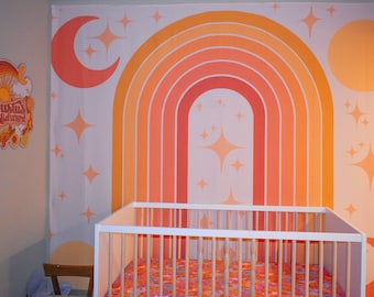 Könnte beinhalten: Ein weißes Babybett mit einem rosa-orangenen Bettuch mit Blumenmuster vor einer Wand mit einem großen orange-rosa-gelben Regenbogenmuster. Das Regenbogenmuster hat einen weißen Hintergrund mit verstreuten Sternen und einem Halbmond auf jeder Seite.