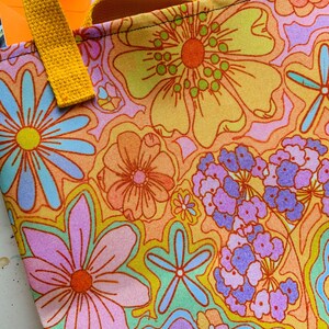 May include: A close-up of a tote bag with a vibrant floral pattern in shades of orange, yellow, pink, and blue. The bag has a yellow handle and a colorful, retro design. The bag is made of fabric.