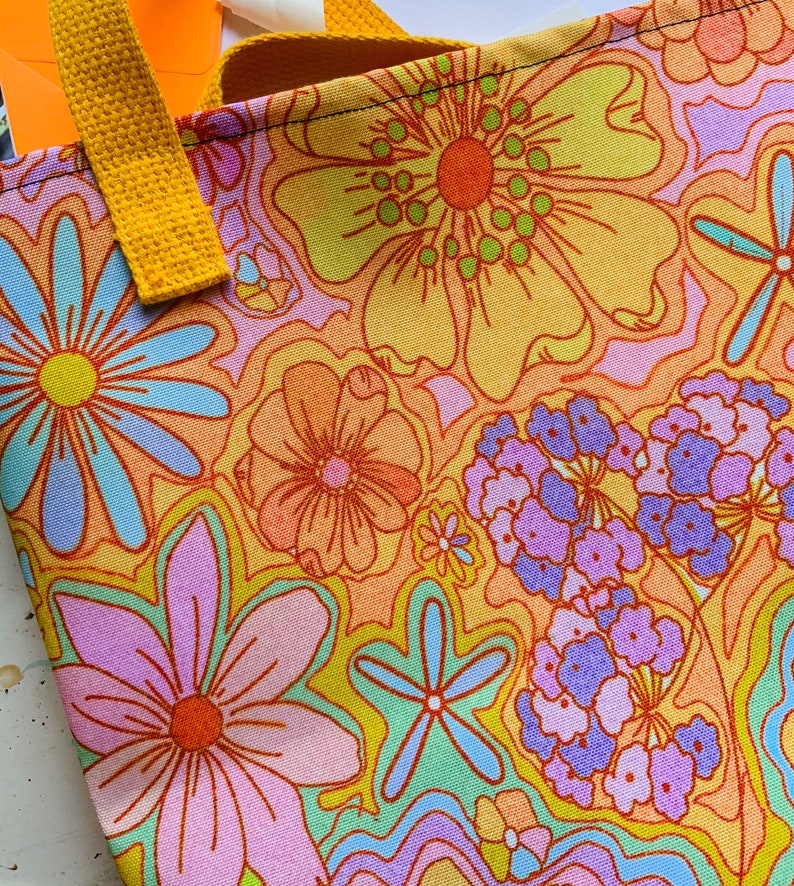 May include: A close-up of a tote bag with a vibrant floral pattern in shades of orange, yellow, pink, and blue. The bag has a yellow handle and a colorful, retro design. The bag is made of fabric.