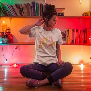 Puede incluir: Una mujer lleva una camiseta blanca con el texto "Going to the party is groovy" en una fuente retro. La camiseta tiene un esquema de color amarillo, naranja y rosa con detalles de purpurina. Est&aacute; sentada en un suelo de madera con las piernas cruzadas.