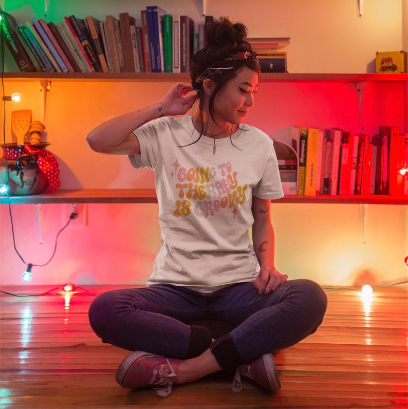 Puede incluir: Una mujer lleva una camiseta blanca con el texto "Going to the party is groovy" en una fuente retro. La camiseta tiene un esquema de color amarillo, naranja y rosa con detalles de purpurina. Est&aacute; sentada en un suelo de madera con las piernas cruzadas.