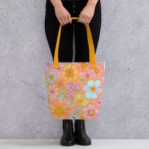May include: A tote bag with a vibrant floral pattern in shades of orange, pink, yellow, and blue. The bag has yellow handles and a rectangular shape, perfect for carrying items. The bag is held by a person.