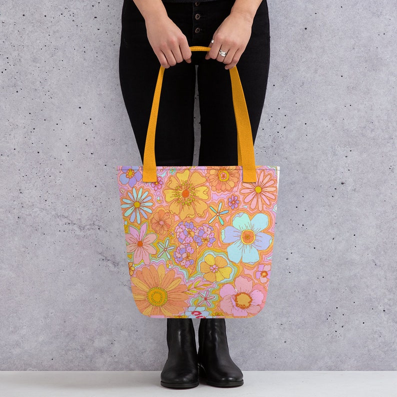 May include: A tote bag with a vibrant floral pattern in shades of orange, pink, yellow, and blue. The bag has yellow handles and a rectangular shape, perfect for carrying items. The bag is held by a person.