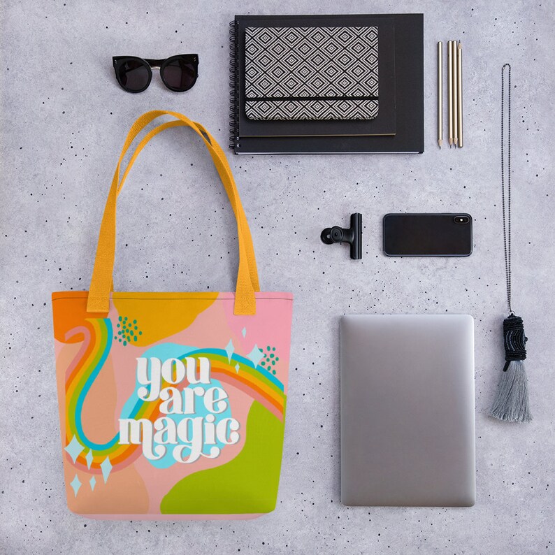 May include: A pink tote bag with the words "you are magic" in white script. The bag features a colorful design with a rainbow, abstract shapes, and yellow handles. Other items include sunglasses, a notebook, pens, a phone, and a laptop.