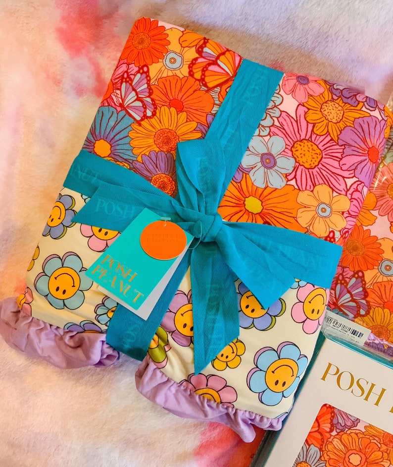 Puede incluir: Una tela blanca con un patr&oacute;n floral y caras sonrientes est&aacute; envuelta en una cinta turquesa y atada con un lazo. La tela est&aacute; etiquetada como "Posh Peanut" y tiene un volante morado alrededor de los bordes.