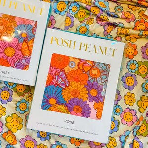 Puede incluir: Una etiqueta de embalaje blanca y azul para una bata con el texto "POSH PEANUT" en letras doradas. La etiqueta presenta una impresi&oacute;n floral colorida con mariposas. El texto "ROBE" est&aacute; impreso debajo de la impresi&oacute;n floral, y el texto "MADE LOVINGLY FROM OUR PAPOOK&trade; VISCOSE FROM BAMBOO" est&aacute; impreso debajo del texto "ROBE".