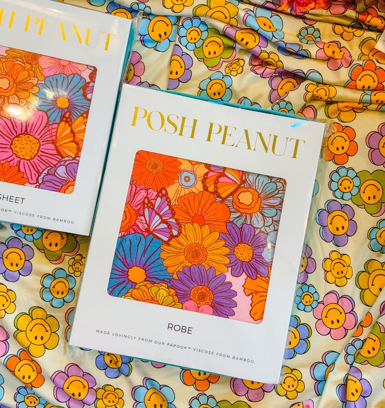 Puede incluir: Una etiqueta de embalaje blanca y azul para una bata con el texto "POSH PEANUT" en letras doradas. La etiqueta presenta una impresi&oacute;n floral colorida con mariposas. El texto "ROBE" est&aacute; impreso debajo de la impresi&oacute;n floral, y el texto "MADE LOVINGLY FROM OUR PAPOOK&trade; VISCOSE FROM BAMBOO" est&aacute; impreso debajo del texto "ROBE".