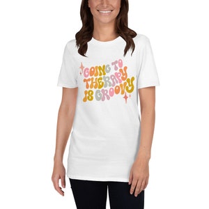 Puede incluir: Camiseta blanca con un gr&aacute;fico de estilo retro que dice "Ir a terapia es genial" en letras coloridas y onduladas.