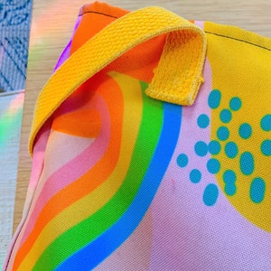May include: A colorful tote bag with a rainbow design. The bag features a vibrant rainbow pattern in shades of orange, yellow, green, and blue. It has a yellow handle and a light pink background with blue polka dots.