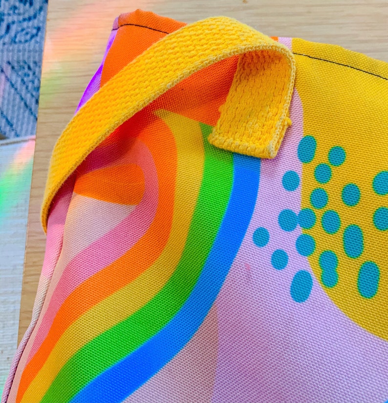 May include: A colorful tote bag with a rainbow design. The bag features a vibrant rainbow pattern in shades of orange, yellow, green, and blue. It has a yellow handle and a light pink background with blue polka dots.