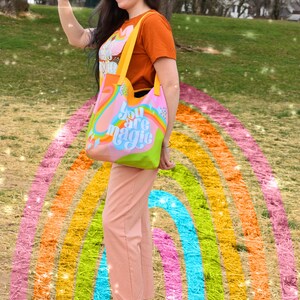 May include: A colorful tote bag with the words "You are magic" in a whimsical font. The bag features a rainbow design with pink, blue, green, and orange hues. The bag has yellow straps and is held by a person wearing an orange t-shirt and pink pants.