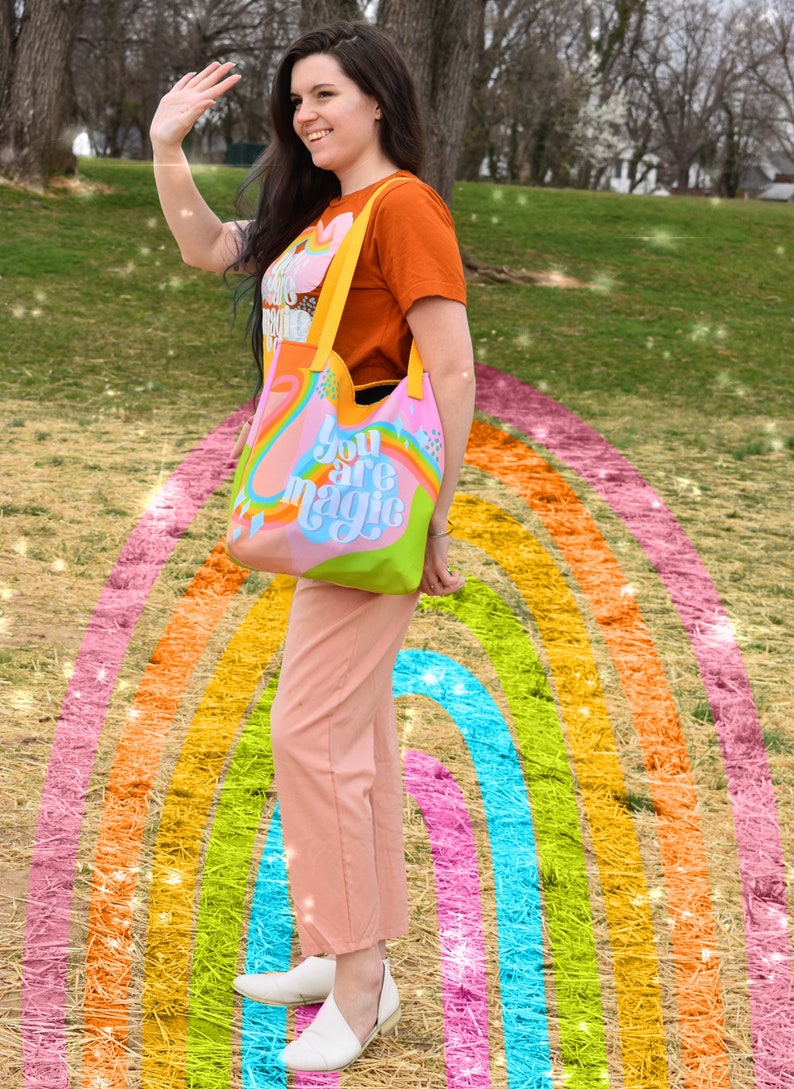 May include: A colorful tote bag with the words "You are magic" in a whimsical font. The bag features a rainbow design with pink, blue, green, and orange hues. The bag has yellow straps and is held by a person wearing an orange t-shirt and pink pants.