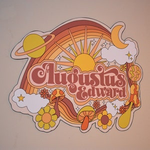 May include: A retro-style graphic with the text "Augustus Edward" in a brown, orange, and yellow color scheme. The design includes a rainbow, sun, planets, clouds, flowers, and mushrooms, evoking a vintage, psychedelic aesthetic.