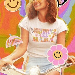 Op de afbeelding: Een vrouw draagt een wit T-shirt met de tekst "Somebody's Groovy Ass Mama" in een retro-lettertype. Ze fietst met een roze bloemen sticker met een smiley erop. De achtergrond is een psychedelische werveling van kleuren.