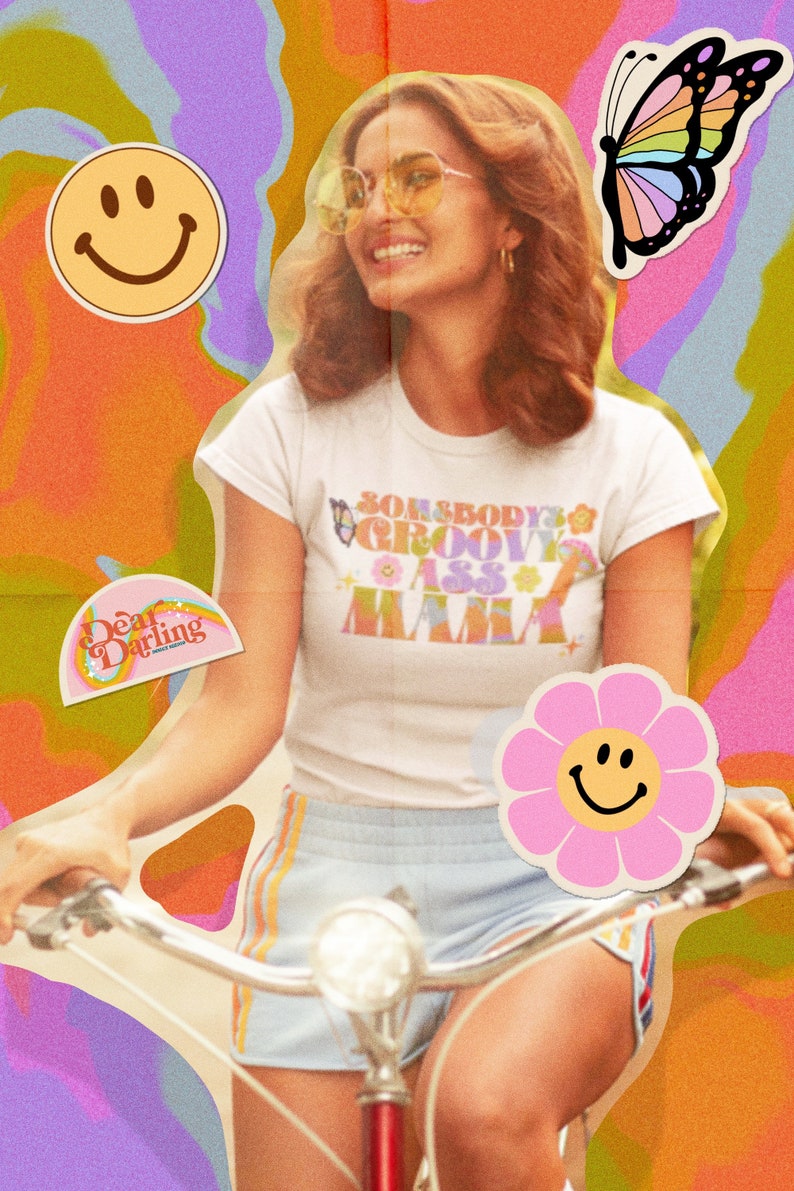 Op de afbeelding: Een vrouw draagt een wit T-shirt met de tekst "Somebody's Groovy Ass Mama" in een retro-lettertype. Ze fietst met een roze bloemen sticker met een smiley erop. De achtergrond is een psychedelische werveling van kleuren.