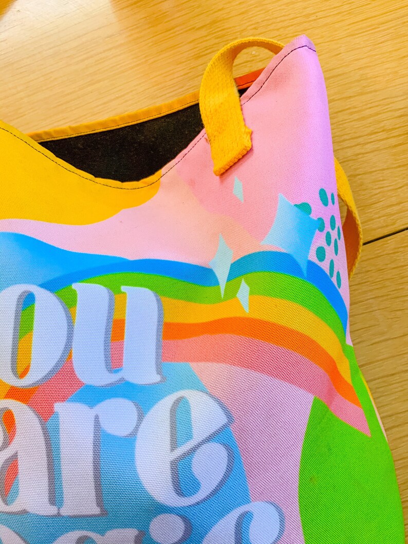 May include: A colorful tote bag with a rainbow design and the words "You are magic" in large white letters. The bag has yellow handles and a pink background with blue and green accents.