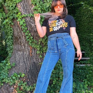 Puede incluir: Una persona lleva una camiseta negra con el texto "Going to therapy is groovy" en colores arco&iacute;ris. Lleva unos vaqueros azules de campana con un dise&ntilde;o de estrellas y un cintur&oacute;n marr&oacute;n.