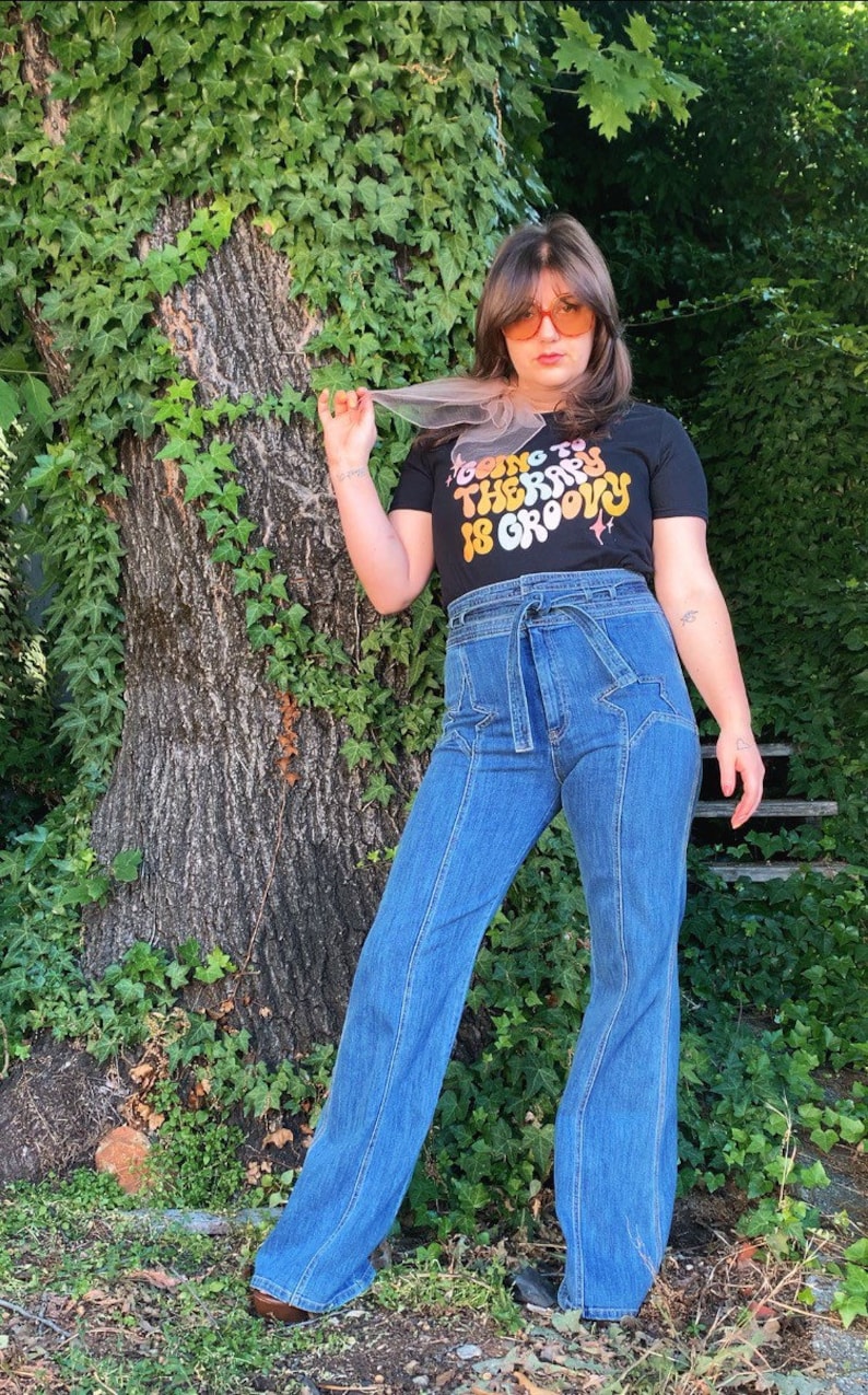 Puede incluir: Una persona lleva una camiseta negra con el texto "Going to therapy is groovy" en colores arco&iacute;ris. Lleva unos vaqueros azules de campana con un dise&ntilde;o de estrellas y un cintur&oacute;n marr&oacute;n.