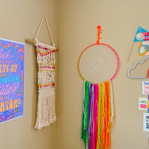 May include: A wall decorated with colourful artwork, including a print with the text "Fuck I feel that getting up in the morning is just creating I am a creator", a macrame wall hanging, a dream catcher with rainbow yarn, a neon cloud with a rainbow, and various other prints.