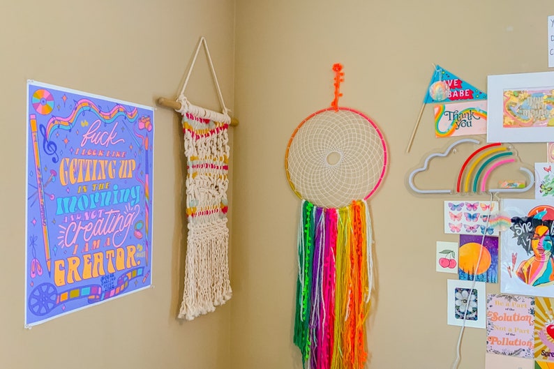 May include: A wall decorated with colourful artwork, including a print with the text "Fuck I feel that getting up in the morning is just creating I am a creator", a macrame wall hanging, a dream catcher with rainbow yarn, a neon cloud with a rainbow, and various other prints.