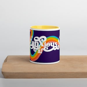 May include: A white ceramic mug with a yellow interior and a dark blue exterior. The mug features the words "anxious af" in white script with a rainbow design. The mug is sitting on a wooden surface.