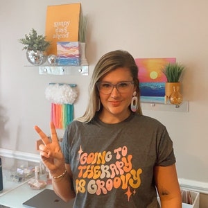 Puede incluir: Una mujer con una camiseta gris con el texto "Going to Therapy is Groovy" en una fuente retro. Lleva pantalones cortos amarillos y tiene el pelo recogido hacia atr&aacute;s. Est&aacute; haciendo un signo de paz con la mano.