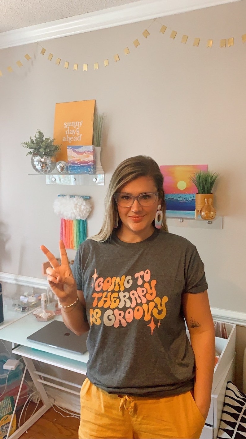 Puede incluir: Una mujer con una camiseta gris con el texto "Going to Therapy is Groovy" en una fuente retro. Lleva pantalones cortos amarillos y tiene el pelo recogido hacia atr&aacute;s. Est&aacute; haciendo un signo de paz con la mano.