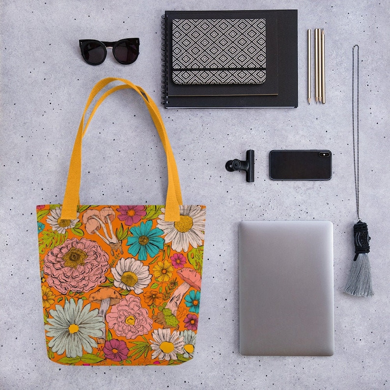 May include: Orange tote bag with a colorful floral and mushroom pattern. The bag has yellow handles and is surrounded by other items, including sunglasses, a notebook, pens, a phone, and a laptop.