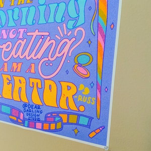 May include: A purple and blue poster with the text "If you're not creating in the morning, you're not a creator." The poster has a colourful design with stars, a film strip, and a makeup brush.