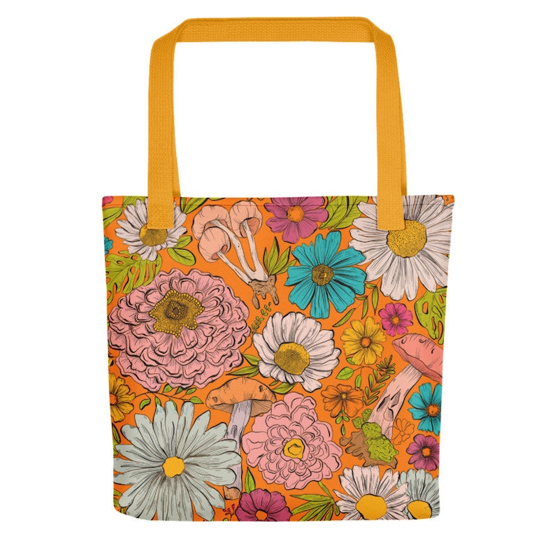 May include: Orange tote bag with a colorful floral and mushroom pattern. The design features various flowers in pink, blue, white, and yellow, along with pink and brown mushrooms. The bag has yellow handles.