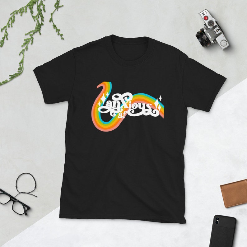 以下が含まれることがあります： レトロな虹のデザインで「Anxious af」の文字が入った黒いTシャツ。デザインは、虹のアーチが付いた、湾曲した様式化されたフォントが特徴です。シャツは柔らかい素材でできており、カジュアルウェアに適しています。