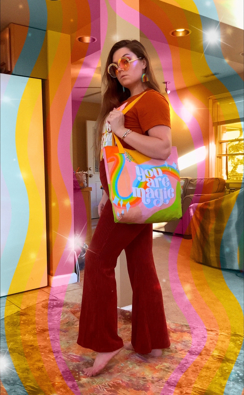 May include: A colorful tote bag with the words "You are magic" in a whimsical font. The bag features a rainbow design and is held by a person wearing a rust-colored top and pants. The background has a retro, psychedelic vibe.