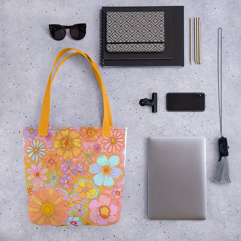 May include: A tote bag with a colorful floral pattern in shades of orange, pink, yellow, and blue. The bag has yellow handles and is surrounded by a black notebook, sunglasses, a phone, and a laptop.