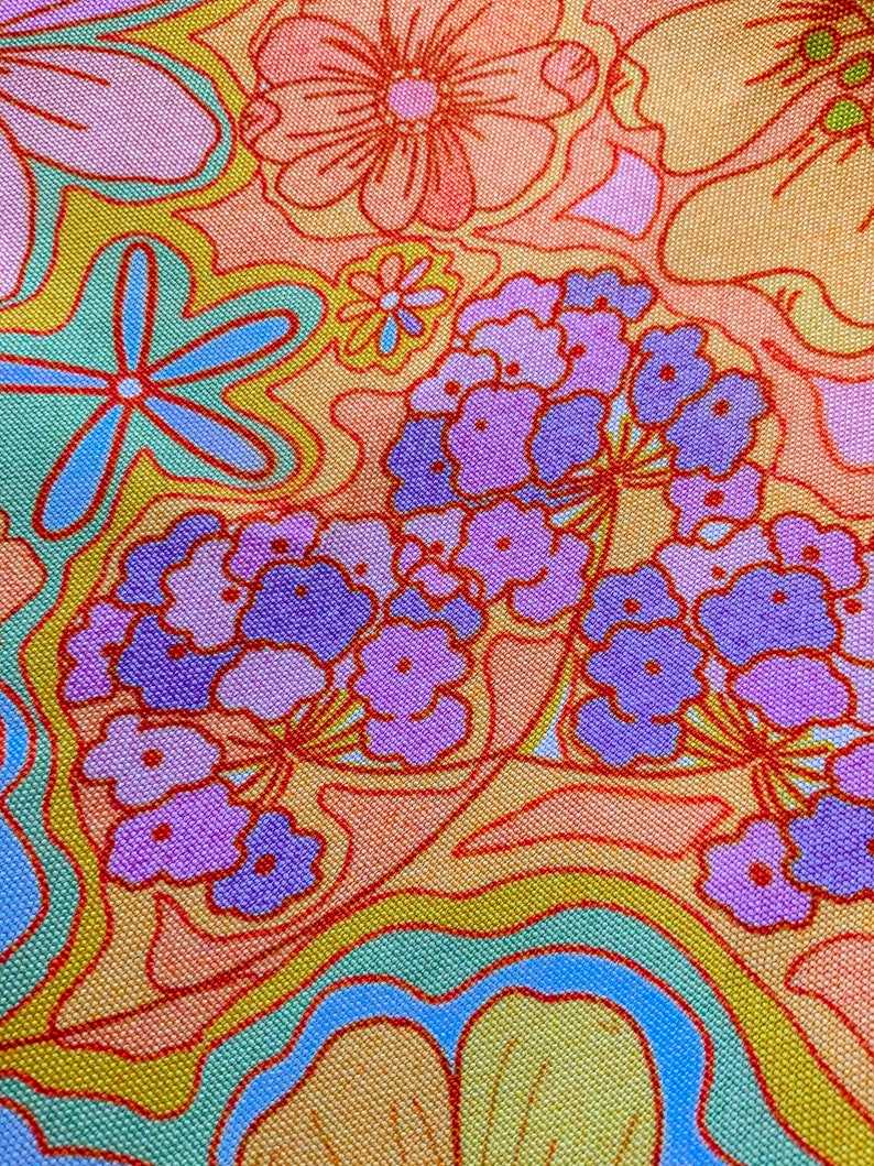 May include: Close-up of a fabric with a vibrant floral pattern. The design features large, stylized flowers in shades of orange, pink, and purple, outlined in red. The background has a mix of yellow, green, and blue wavy lines.