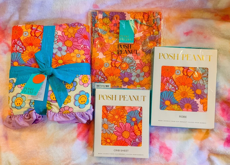 Puede incluir: Tres paquetes de ropa de cama y ropa con un estampado floral retro con mariposas y caritas sonrientes. Los paquetes est&aacute;n etiquetados como "Posh Peanut" e incluyen una s&aacute;bana de cuna, una bata y una manta.