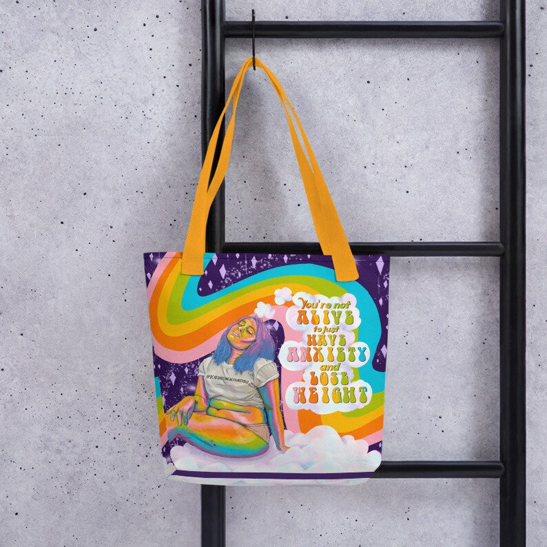 May include: A tote bag with a vibrant design featuring a seated figure with blue hair, a rainbow, and the text "You're not alive to just have anxiety and lose height." The bag has orange handles and a purple background.