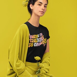 Puede incluir: Una mujer con una camiseta negra con el texto "Going to therapy is groovy" en una fuente retro. Lleva un c&aacute;rdigan y una falda amarillos y tiene flores amarillas en el pelo.