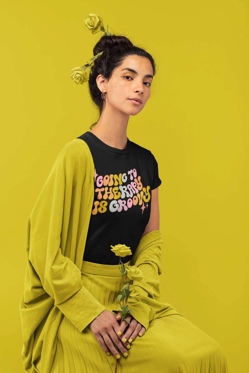 Puede incluir: Una mujer con una camiseta negra con el texto "Going to therapy is groovy" en una fuente retro. Lleva un c&aacute;rdigan y una falda amarillos y tiene flores amarillas en el pelo.