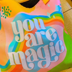 May include: A colorful tote bag with the words "you are magic" in white block letters. The bag features a vibrant design with rainbow stripes, blue, orange, and green hues. It has a yellow handle and a black interior.