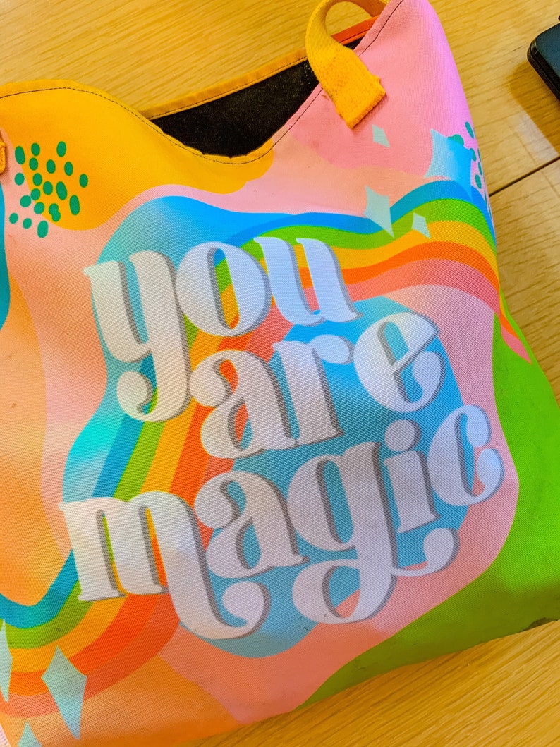 May include: A colorful tote bag with the words "you are magic" in white block letters. The bag features a vibrant design with rainbow stripes, blue, orange, and green hues. It has a yellow handle and a black interior.