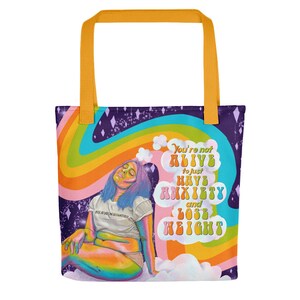 May include: A tote bag with a vibrant design featuring a rainbow, clouds, and a figure with blue and purple hair. The bag has yellow handles and text that reads "You're not ALIVE to just HAVE ANXIETY and LOSE WEIGHT."