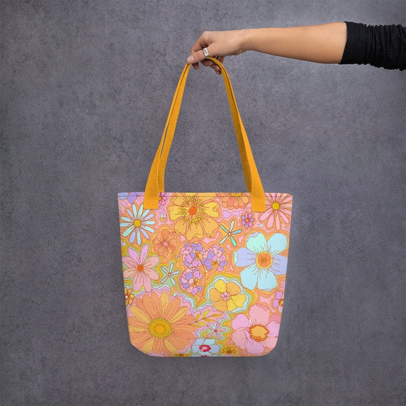 May include: A tote bag with a colorful floral pattern in shades of orange, pink, yellow, and blue. The bag has mustard yellow handles and is held up against a gray background. The floral design is reminiscent of the 1960s and 1970s.