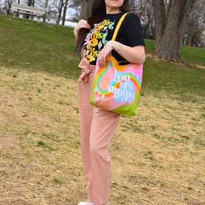May include: A black t-shirt with colorful text that reads "The artist is this". A tote bag with the words "You are magic" in a rainbow design. The person is wearing pink pants and white shoes. The background is a park with trees and grass.