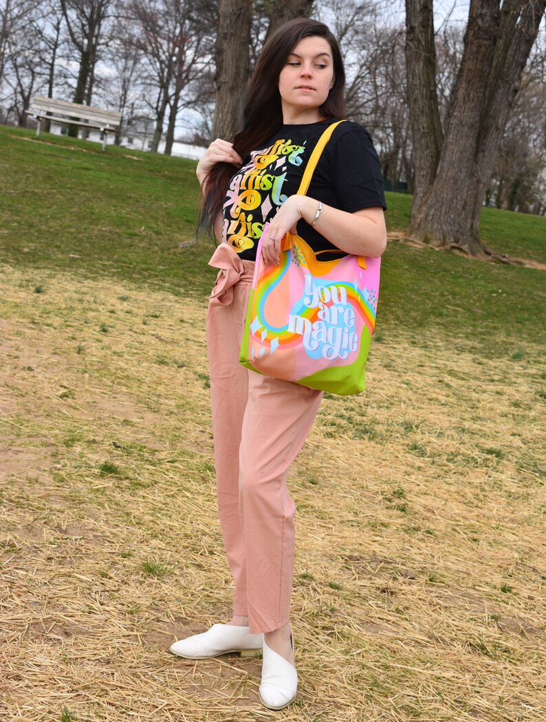 May include: A black t-shirt with colorful text that reads "The artist is this". A tote bag with the words "You are magic" in a rainbow design. The person is wearing pink pants and white shoes. The background is a park with trees and grass.