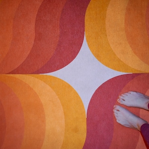 May include: A retro-style rug with a repeating pattern of orange, yellow, and red semi-circles. The design creates a circular pattern. The image also shows a person's feet and legs wearing purple leggings.