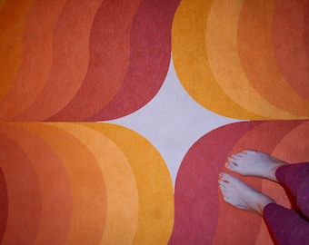 Könnte beinhalten: Ein Teppich im Retro-Stil mit einem sich wiederholenden Muster aus orangefarbenen, gelben und roten Halbkreisen. Das Design erzeugt ein kreisförmiges Muster. Das Bild zeigt auch die Füße und Beine einer Person, die lila Leggings trägt.