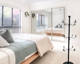May include: A black metal candle holder with multiple arms for candles. The holder is on a round base and stands on a white floor. The holder is in a bedroom with a bed and a mirror in the background.